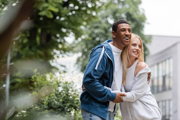 Joyeux couple interracial en sweat à capuche embrassant et regardant loin sur la rue verte et floue — Photo de stock