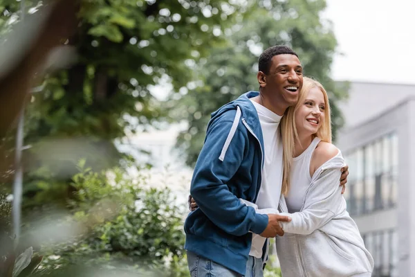 Freudiges gemischtrassiges Paar in Kapuzenpullis, das sich auf der grünen und verschwommenen Straße umarmt und wegsieht — Stockfoto