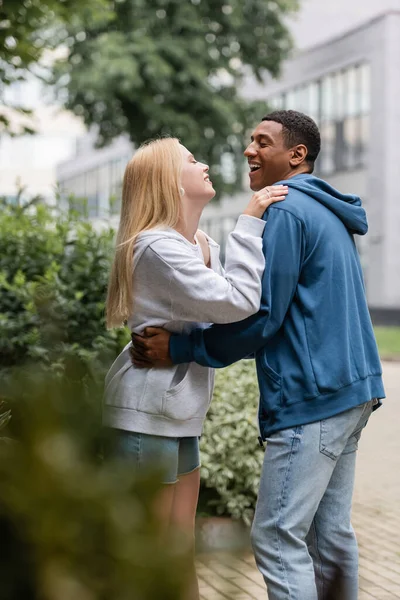 Vista lateral do homem afro-americano rindo e mulher loira abraçando e olhando um para o outro na rua — Fotografia de Stock
