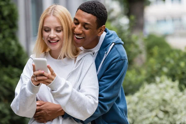 Улыбающийся африканский американец обнимает блондинку, обмениваясь сообщениями со смартфоном на улице — Stock Photo