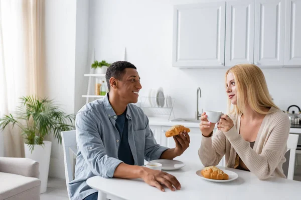 Glückliches interrassisches Paar, das sich beim Frühstück mit Kaffee und leckeren Croissants anschaut — Stockfoto