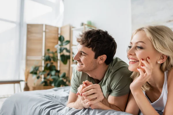Happy man in pajama holding hand of blonde girlfriend on bed in morning — Stock Photo