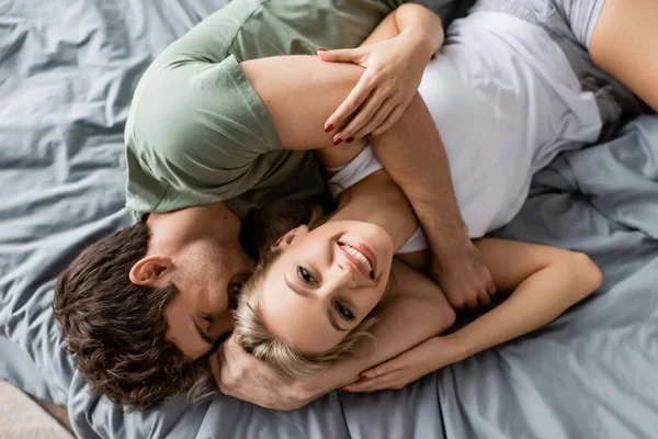 Top view of cheerful man hugging girlfriend in pajama looking at camera on bed — Stock Photo
