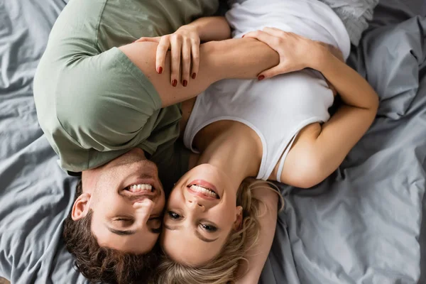 Vista dall'alto di allegra giovane coppia in pigiama guardando la fotocamera mentre si abbraccia sul letto — Foto stock