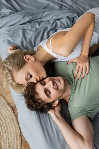 Vue du dessus de la jeune femme embrassant petit ami joyeux en pyjama sur le lit à la maison — Photo de stock