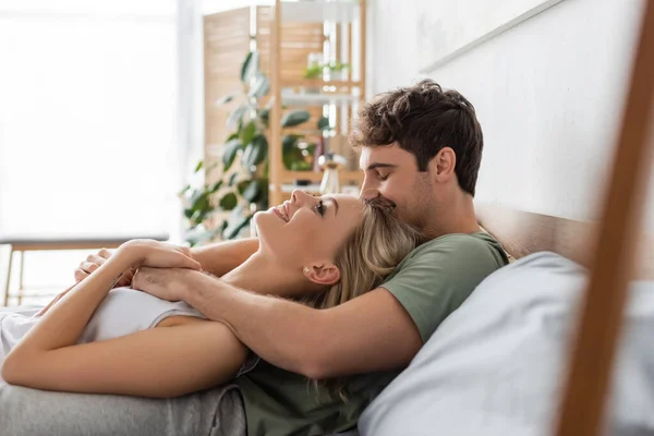 Vista lateral de un hombre joven besando a su novia rubia en la cama por la mañana - foto de stock