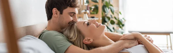 Seitenansicht eines lächelnden Mannes, der morgens seine hübsche blonde Freundin auf dem Bett umarmt, Banner — Stockfoto