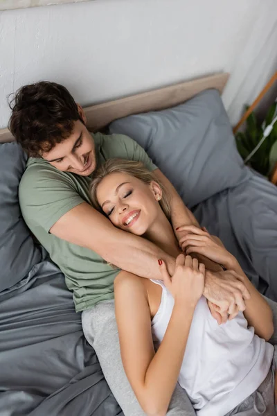 Vista ad alto angolo di uomo sorridente in pigiama abbracciare la ragazza soddisfatta sul letto — Foto stock