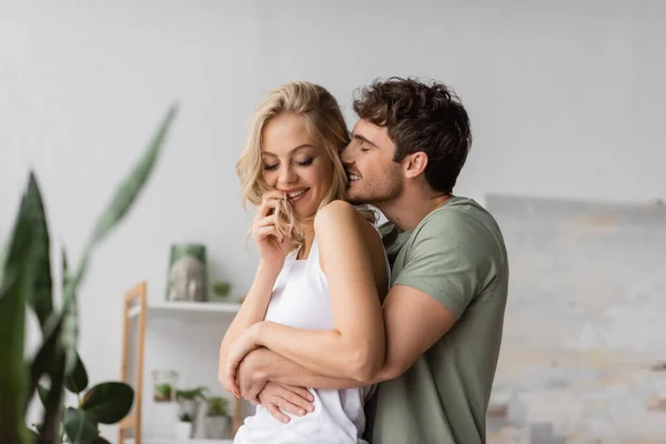 Молодой человек обнимает и целует веселую девушку в пижаме дома — стоковое фото