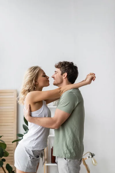 Side view of blonde woman in pajama kissing and embracing boyfriend at home — Stock Photo