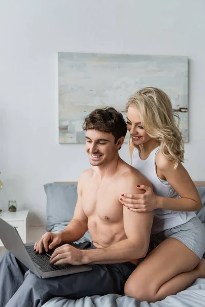 Mujer rubia alegre abrazando novio muscular con portátil en la cama - foto de stock