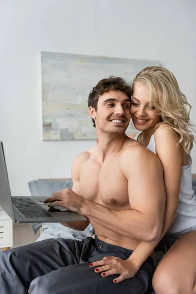 Гарненька блондинка торкається м'язистого хлопця з ноутбуком в спальні — стокове фото
