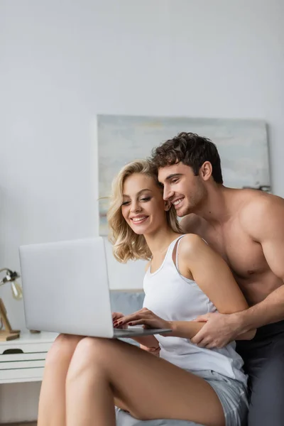Shirtless man hugging blonde girlfriend with laptop in bedroom — Stock Photo