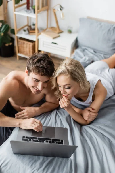 Vue grand angle de sourire homme torse nu en utilisant un ordinateur portable près de la petite amie en pyjama sur le lit — Photo de stock