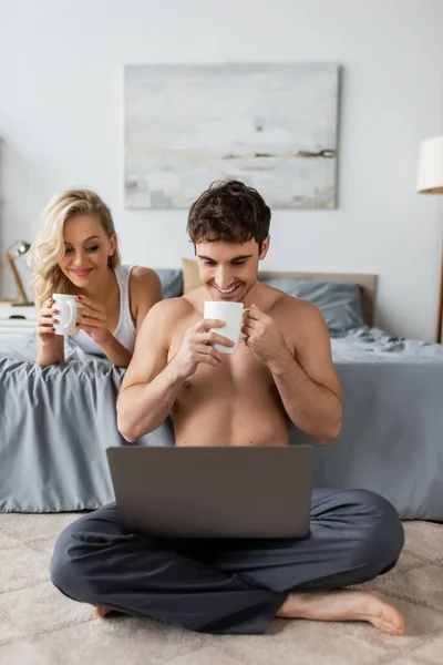 Positivo hombre musculoso sosteniendo la taza de café y mirando a la computadora portátil cerca de la novia en la cama por la mañana - foto de stock