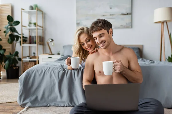 Pareja sonriente con tazas de café mirando a la computadora portátil en el dormitorio por la mañana - foto de stock