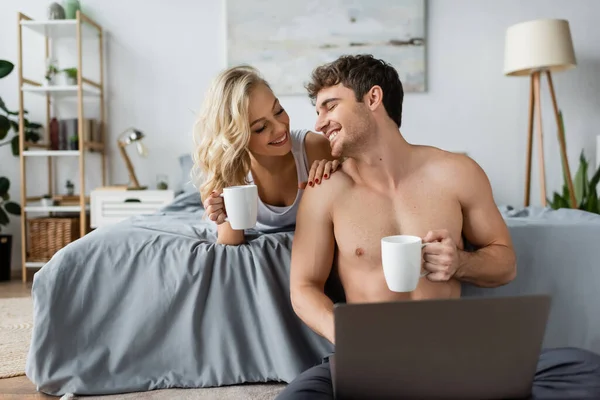 Femme positive en pyjama tenant une tasse de café et touchant petit ami musclé avec ordinateur portable dans la chambre — Photo de stock