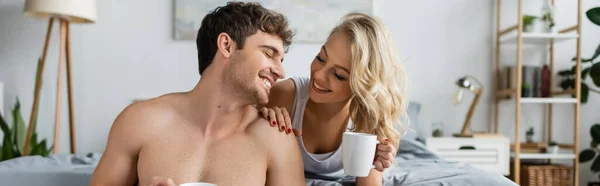 Mulher loira alegre segurando xícara de café perto de namorado sem camisa no quarto, banner — Fotografia de Stock