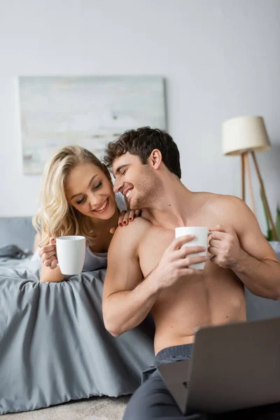 Sonriente hombre sin camisa sosteniendo taza de café cerca de la novia rubia y el ordenador portátil en el dormitorio - foto de stock