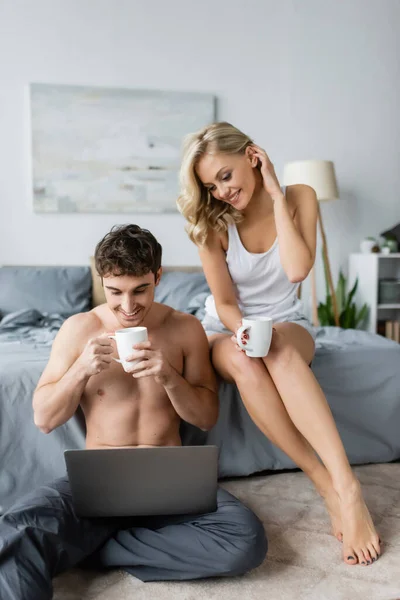 Femme blonde souriante en pyjama tenant tasse de café près de copain torse nu avec ordinateur portable dans la chambre — Photo de stock