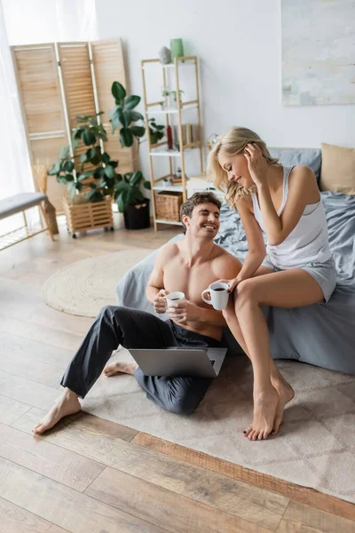 Fröhliches sexy Paar hält Kaffee in der Nähe von Laptop im Schlafzimmer — Stockfoto