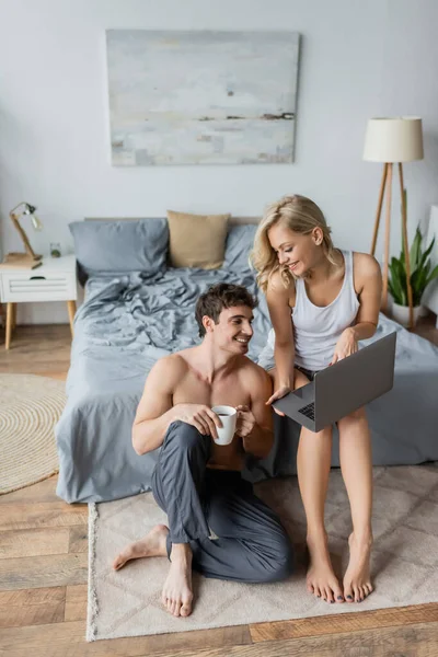 Smiling woman in pajama holding laptop near sexy boyfriend with coffee in bedroom — Stock Photo
