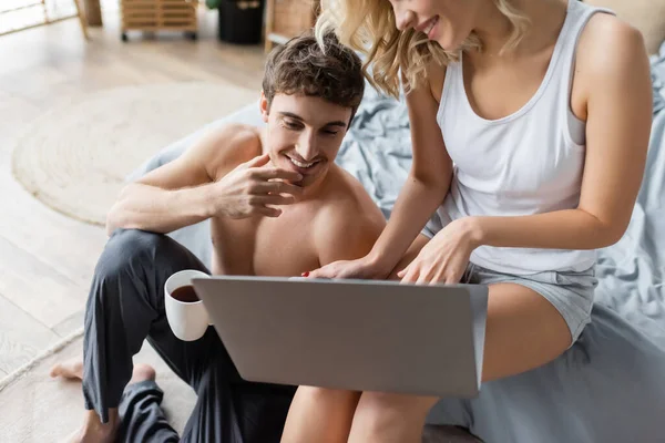 Sorrindo mulher loira no pijama segurando laptop perto namorado sexy com café no quarto — Fotografia de Stock