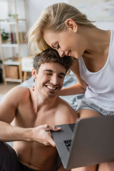 Mujer rubia sonriente en pijama abrazando novio sin camisa con portátil en el dormitorio - foto de stock