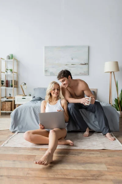 Улыбающийся мужчина без рубашки держит чашку кофе рядом с подругой с ноутбуком в спальне — стоковое фото