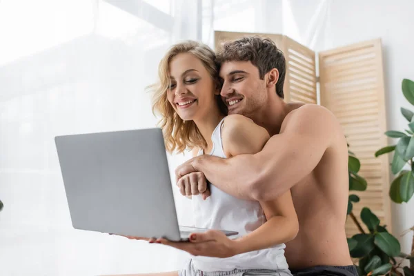 Muscular man hugging girlfriend in pajama holding laptop at home — Stock Photo