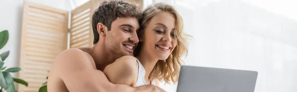 Без сорочки чоловік обіймає усміхнену дівчину біля ноутбука вдома, банер — стокове фото