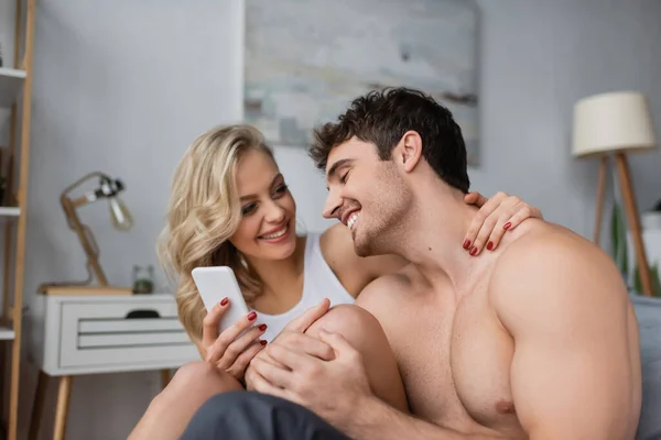 Mujer rubia sonriente sosteniendo teléfono inteligente y abrazando novio sin camisa en el dormitorio - foto de stock