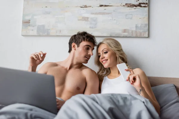 Улыбающаяся блондинка держит смартфон рядом с сексуальным парнем с ноутбуком на кровати — стоковое фото