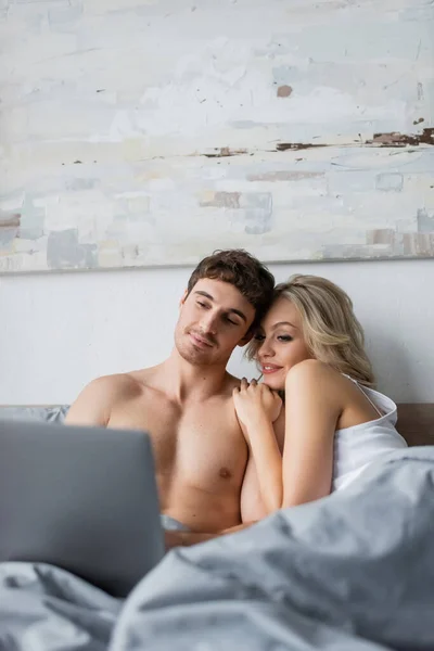 Blonde woman hugging sexy boyfriend and looking at blurred laptop on bed — Stock Photo