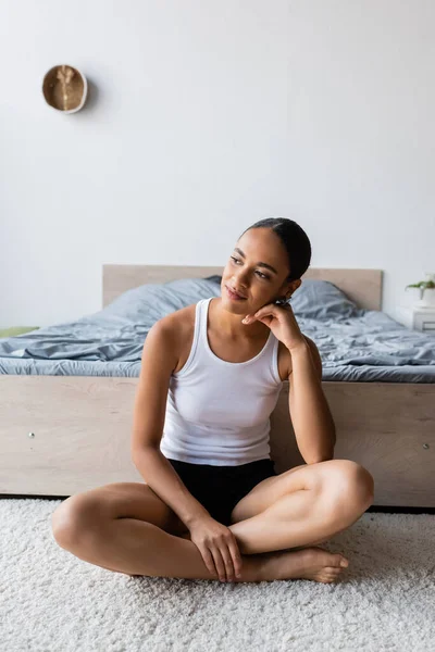 Joven y soñadora mujer afroamericana en camiseta sin mangas sentada cerca de la cama - foto de stock