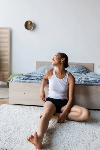 Young and cheerful african american woman in tank top sitting near bed — Stock Photo