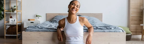 Joven y alegre mujer afroamericana en camiseta sin mangas sentada cerca de la cama, pancarta - foto de stock