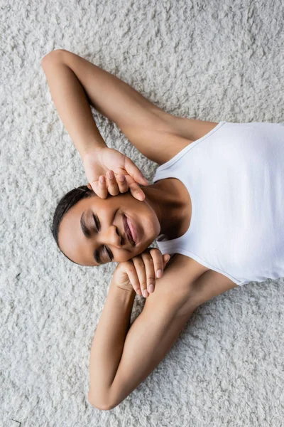 Vista superior de la mujer afroamericana complacida en camiseta sin mangas acostada en la alfombra - foto de stock