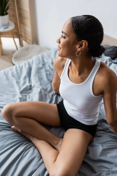 Vista de ángulo alto de la mujer afroamericana complacida en camiseta sin mangas y pantalones cortos sentados en la cama - foto de stock