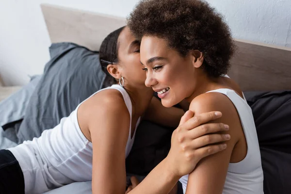 Africano americano lesbiana mujer besos mejilla de feliz novia en dormitorio - foto de stock