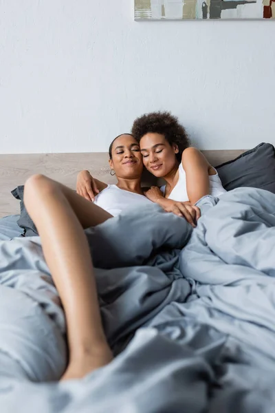 Happy african american same sex couple with closed eyes lying on bed at home — Stock Photo