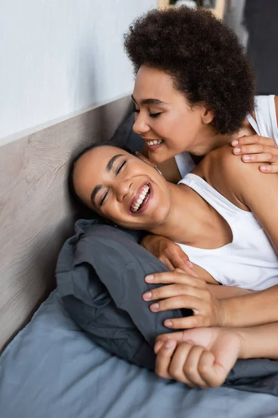 Joyful and lesbian african american woman with closed eyes hugging girlfriend on bed — Stock Photo
