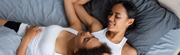 Вид сверху на кудрявую африканскую лесбиянку, лежащую со счастливой подругой на кровати, баннер — стоковое фото