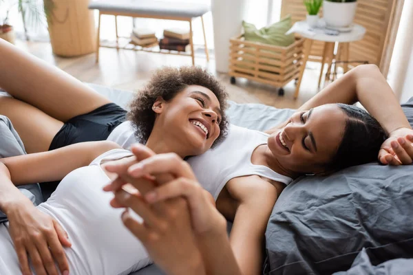 Alegre lesbiana africana americana mujer cogida de la mano mientras se acuesta con feliz novia en la cama - foto de stock