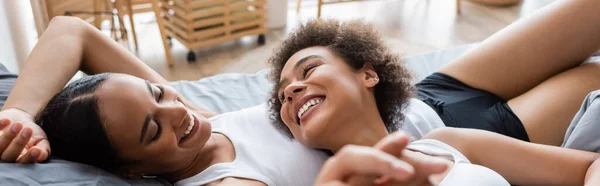 Lesbiana afroamericana americana mujer cogida de la mano mientras se acuesta con feliz novia en la cama, pancarta - foto de stock