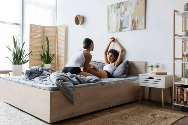 Young lesbian african american woman looking at happy girlfriend with raised hands in modern bedroom — Stock Photo