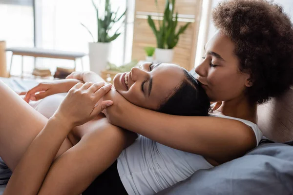 Lesbiana africana americana mujer besos cabeza de complacido novia en moderno dormitorio - foto de stock
