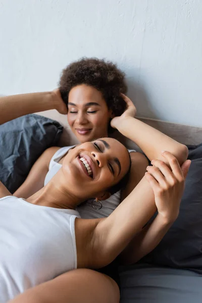 Lesbiana africana americana mujer abrazando cabeza de complacido novia en moderno dormitorio - foto de stock
