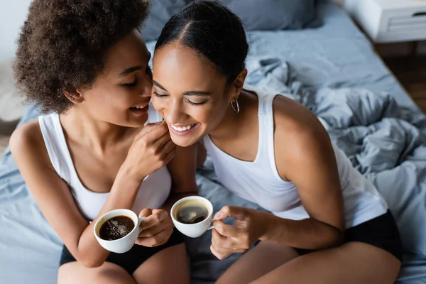 Высокий угол зрения счастливой лесбиянки африканской американской пары держа чашки кофе, сидя на кровати — стоковое фото