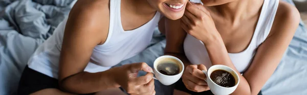 Обрезанный вид на счастливую лесбийскую африканскую пару, держащую чашки кофе в спальне, баннер — стоковое фото